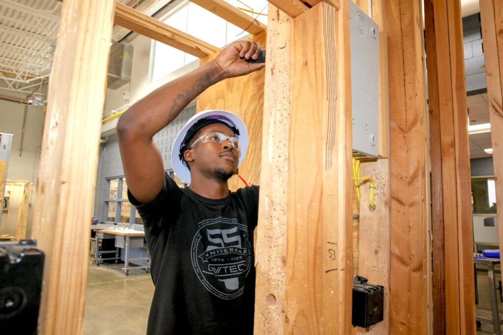 Student with hard hat and safety glasses on working on electrical wiring for structure frame.