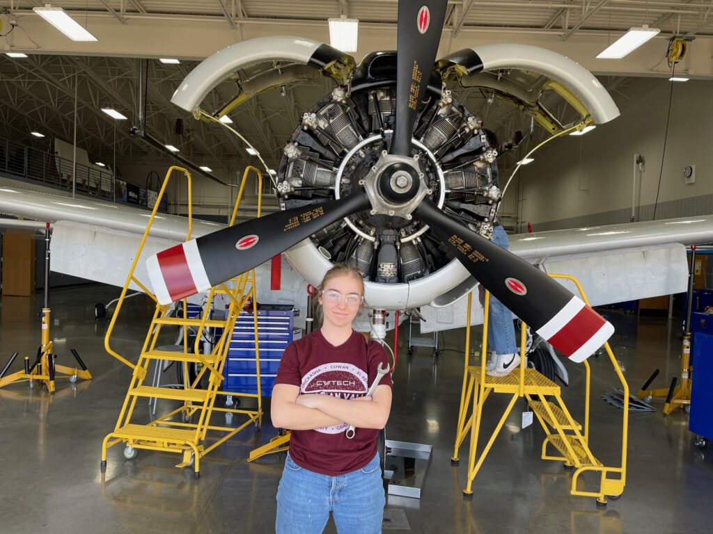 Young adult female stands in front of a single-prop aircraft while holding a wrench.