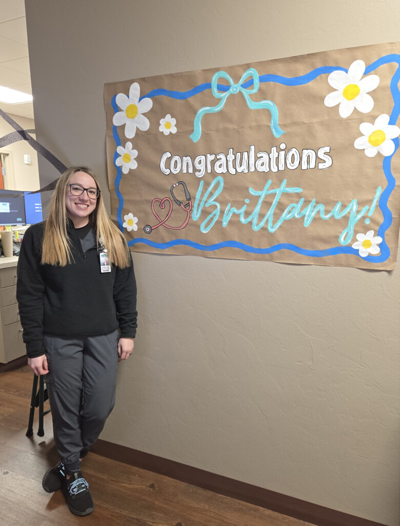 Young lady in late 20s stands beside a wall banner that reads, "Congratulations Brittany!" and features a cartoon stethoscope and various sizes of daisies.