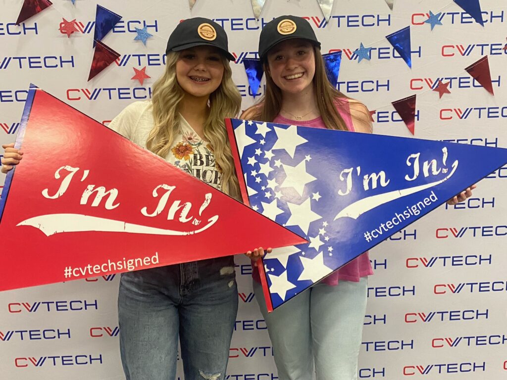 Two gils in hats hold giant cardboard pennants in front of a CV Tech backdrop with red and blue star garland.