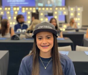 smiling student sits at a table wearing a CV Tech hat with a large crowd behind her.