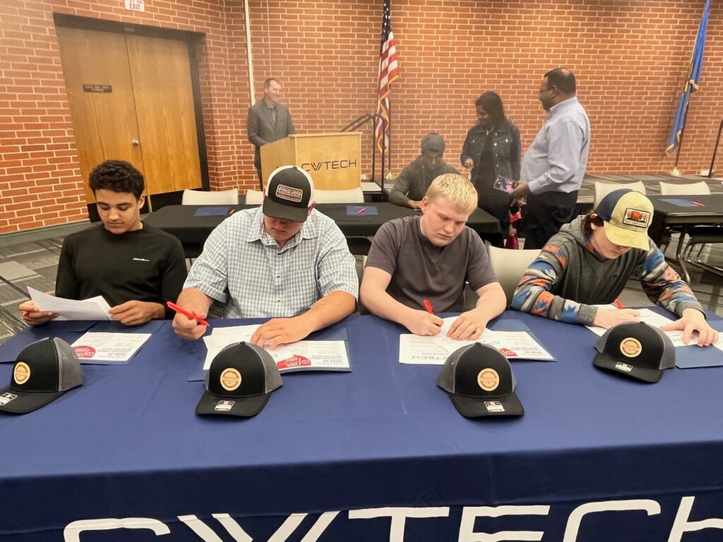 4 boys sit at a CV Tech table and sign paperwork with new hats in front of them.
