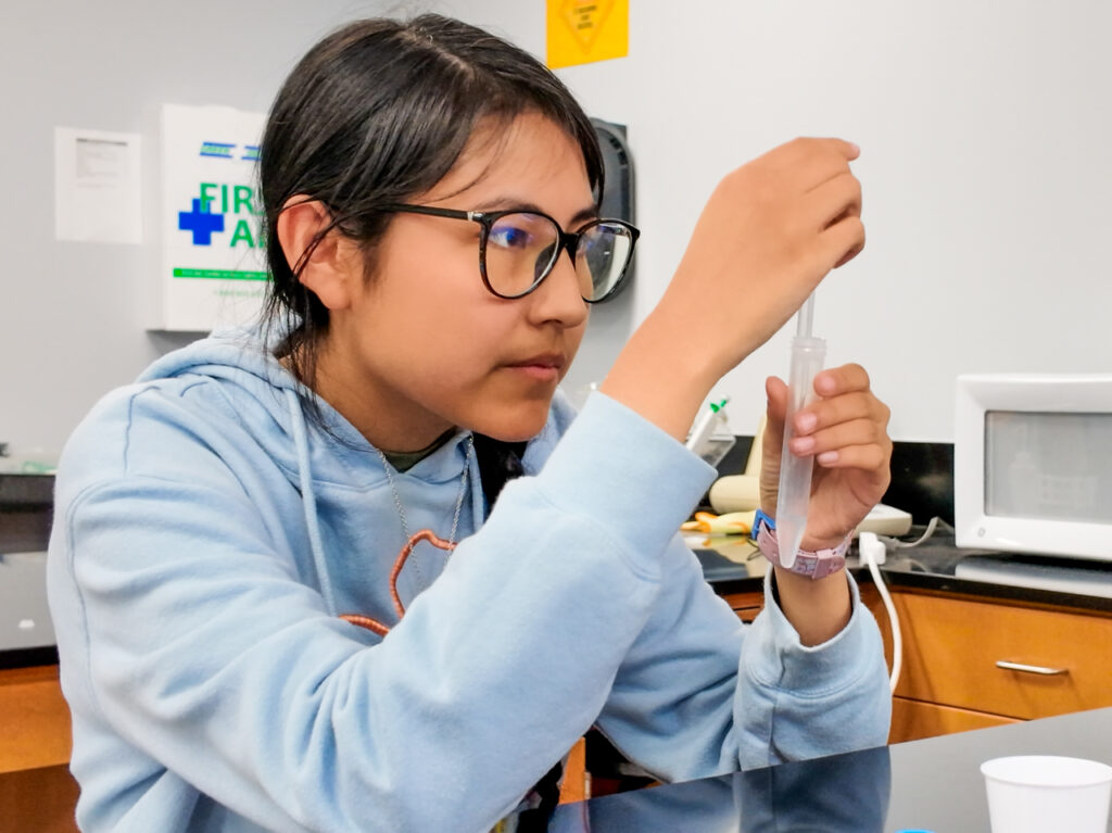 A high school student with glasses stares intently at a test tube being held in her hands.