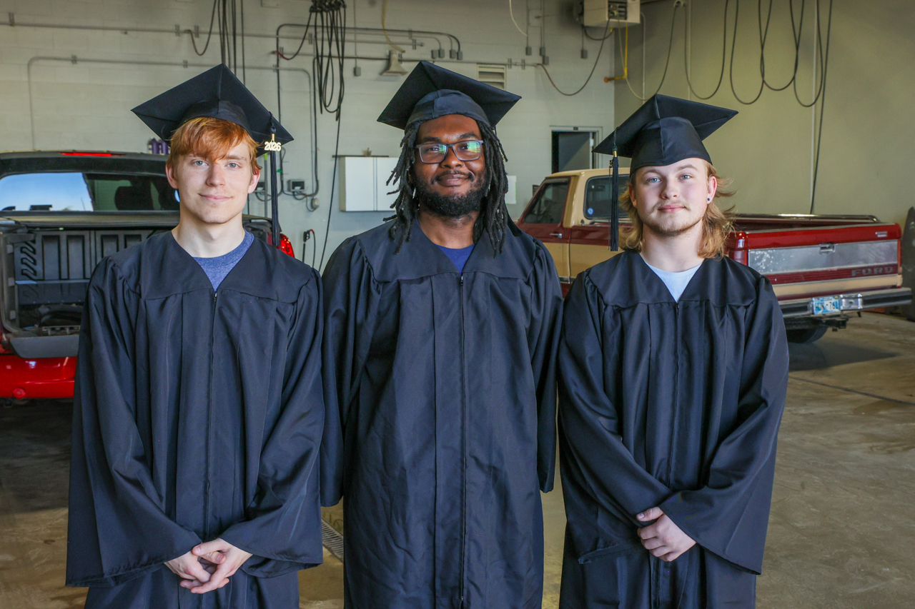 three students n caps and gowns stand in front of trucks in an auto shop setting.