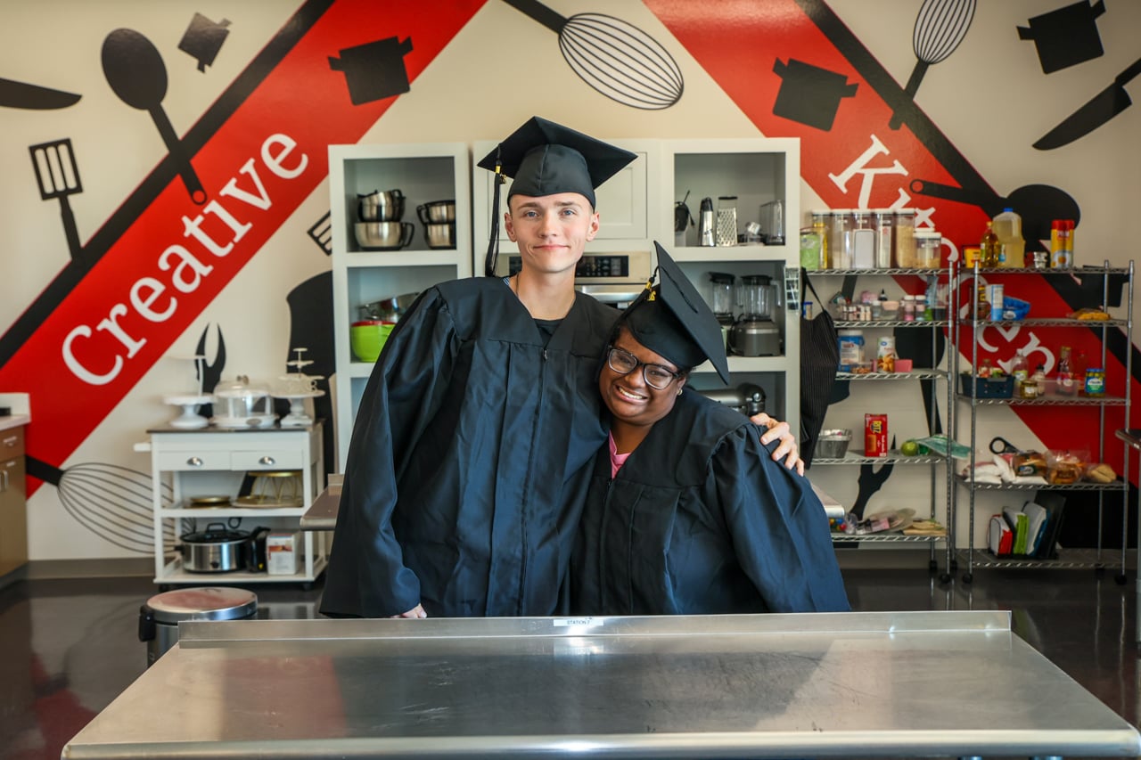 Two students in caps and gowns smile and lean into each other