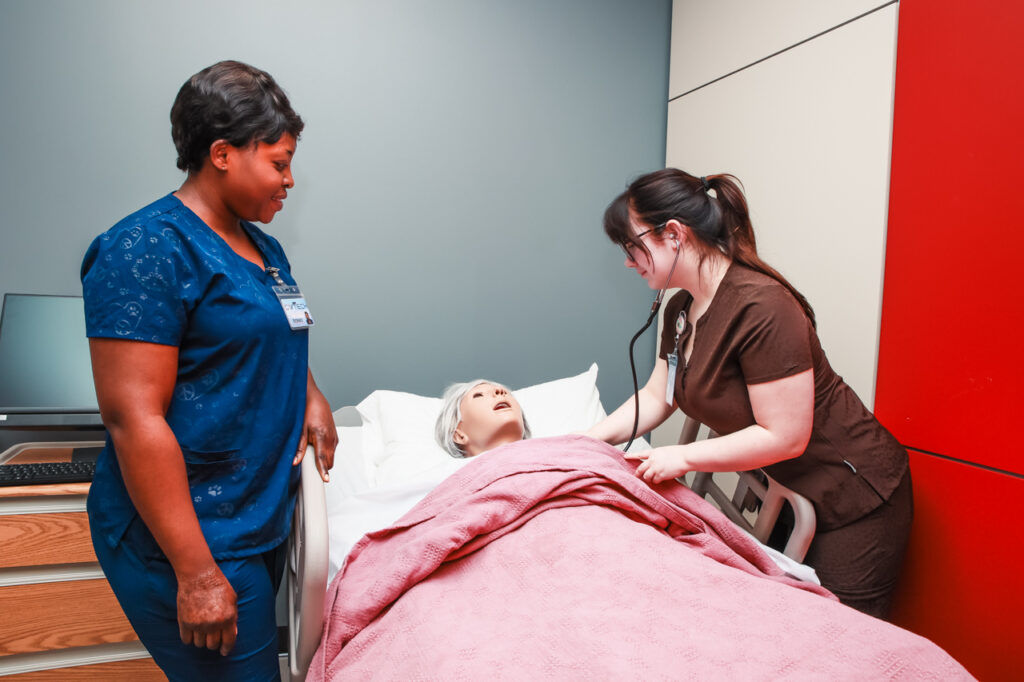 Two students in scrubs stand beside a hospital bed on either side of an elderly female mannequin with gray hair.