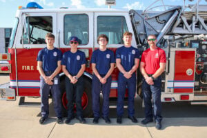 Four students in matching navy T-shirts stand next to their instructor and in front of a fire engine.