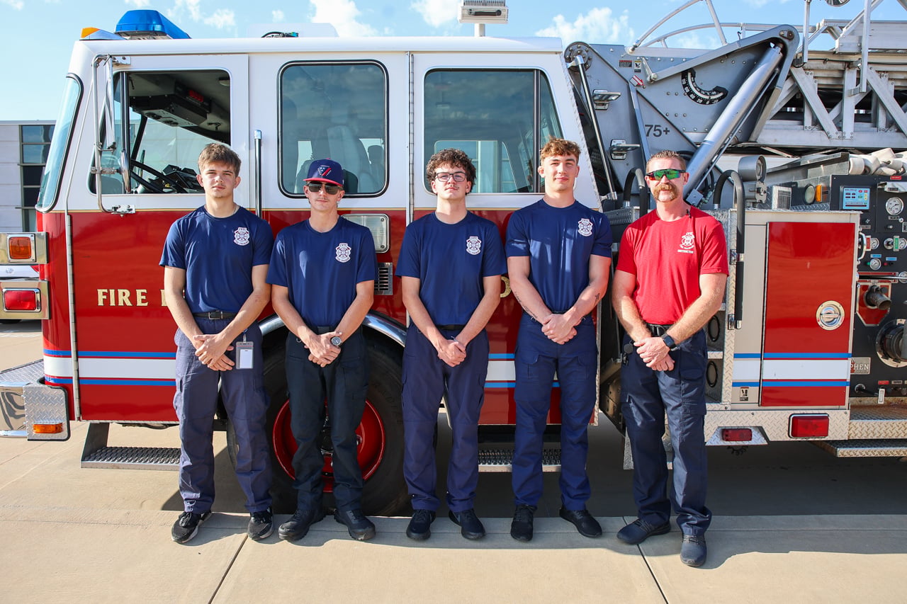 Four students in matching navy T-shirts stand next to their instructor and in front of a fire engine.