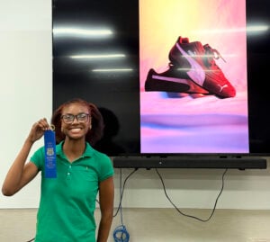 A student holds the blue ribbon she won at the recent Oklahoma State Fair for a photograph of red Puma shoes set on a backdrop with yellow, orange and purple hues.