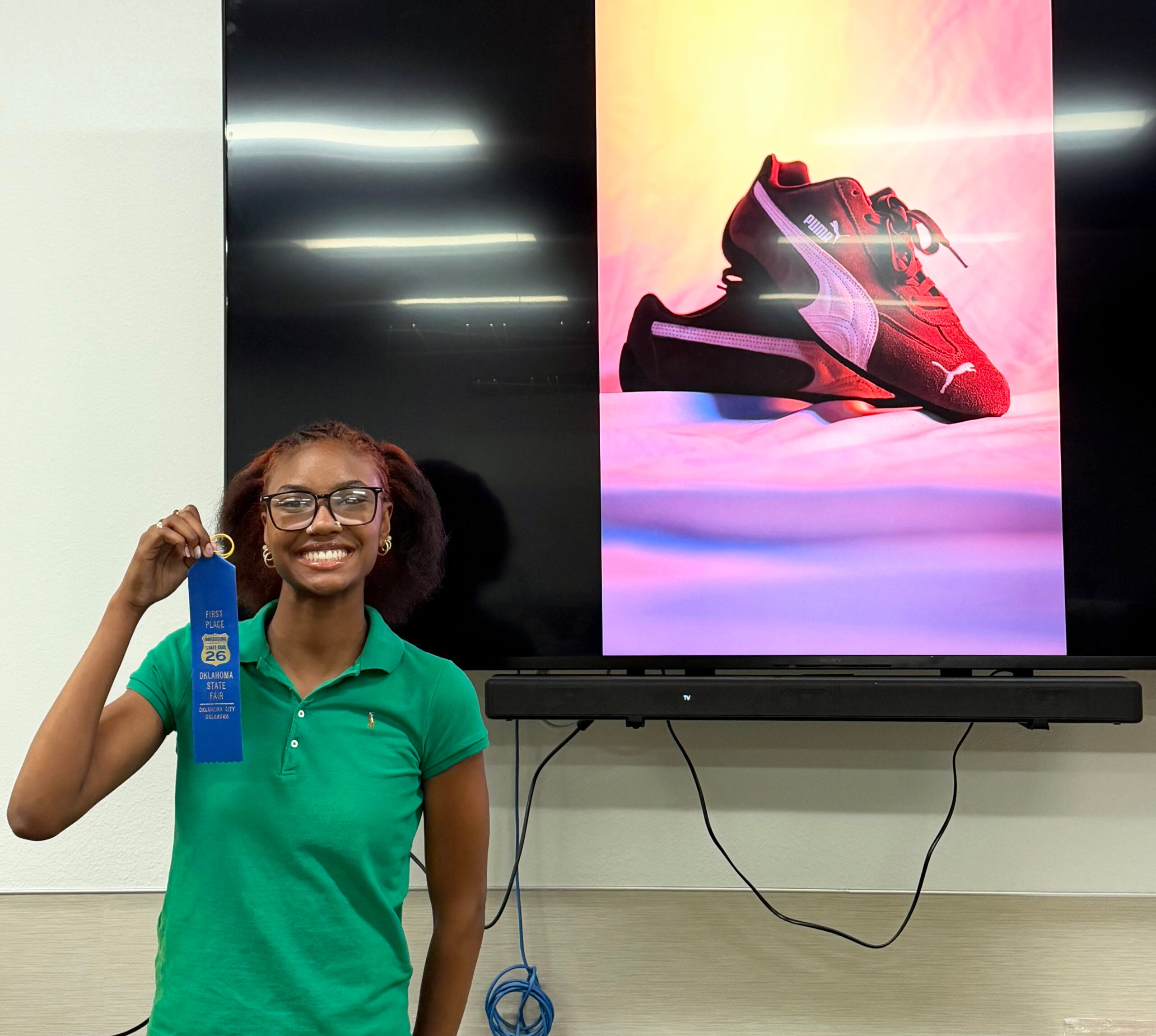 A student holds the blue ribbon she won at the recent Oklahoma State Fair for a photograph of red Puma shoes set on a backdrop with yellow, orange and purple hues.