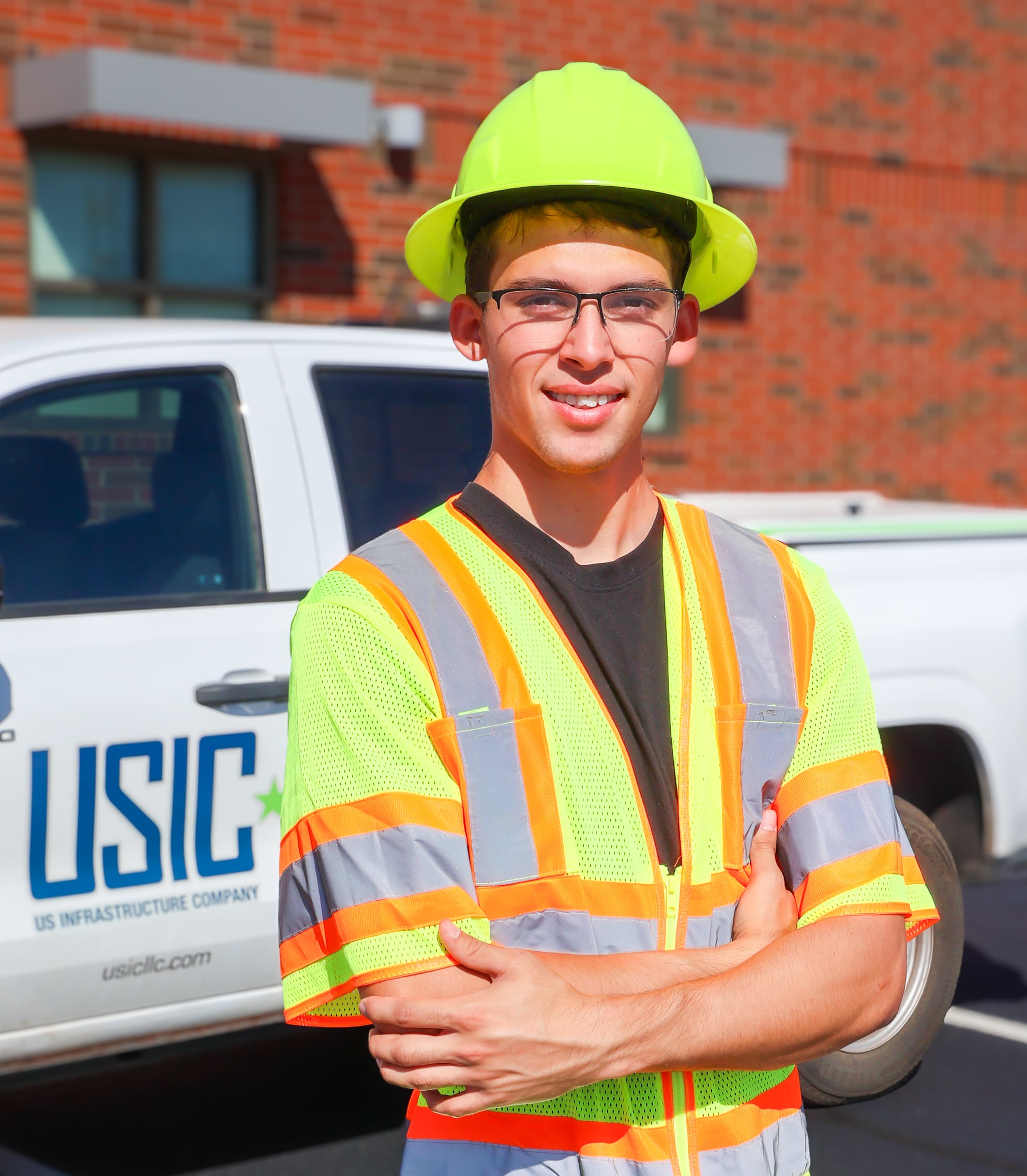 A young man is dressed in a bright green hard hat and vest, standing in front of a pickup with the letters "USIC" on the door.