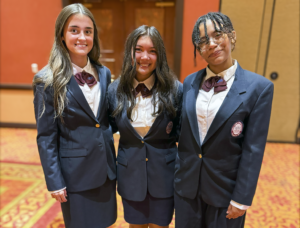 Three young ladies, dressed in navy suit jackets and maroon crossover tie are smiling while posing for a photo.