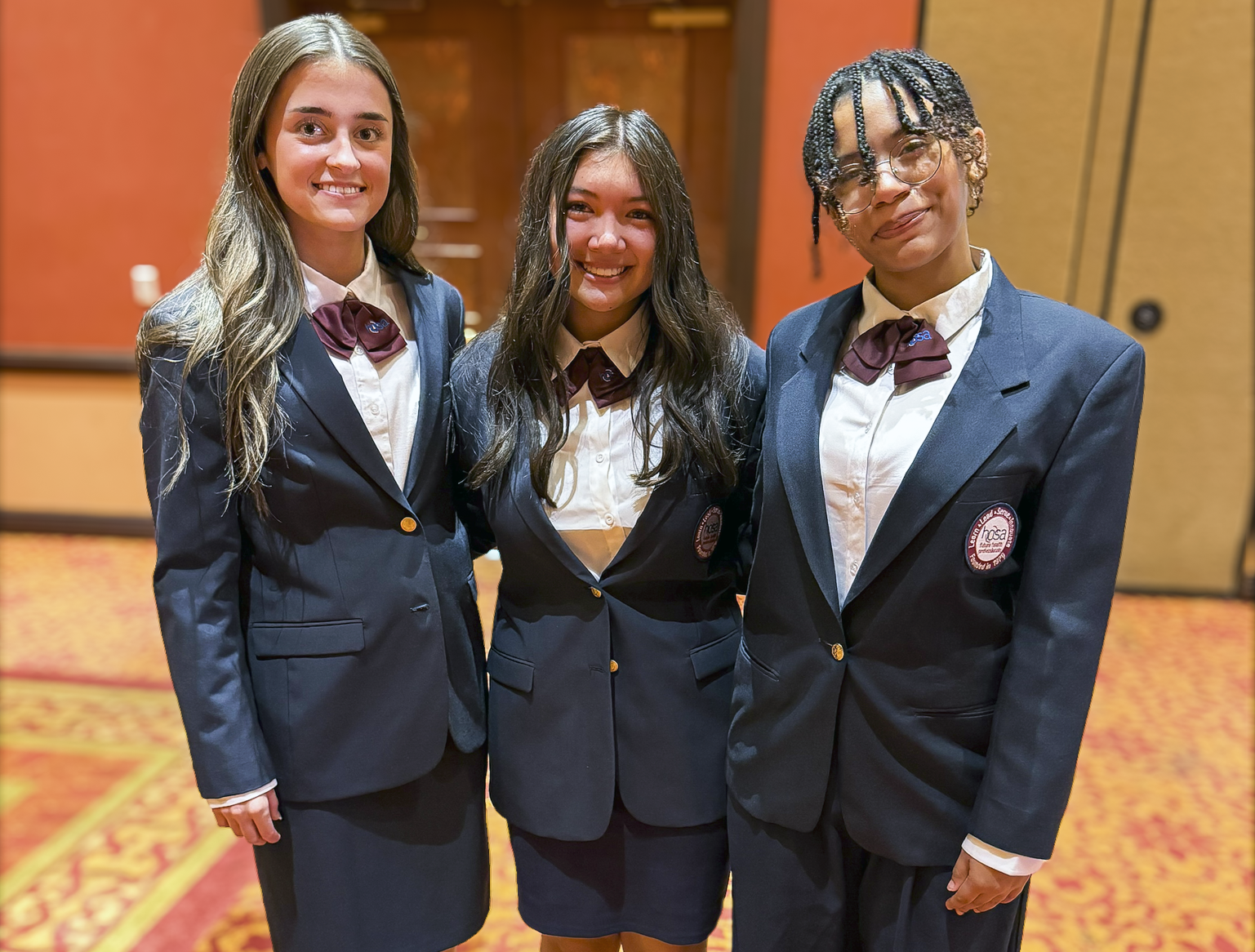 Three young ladies, dressed in navy suit jackets and maroon crossover tie are smiling while posing for a photo.